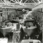 View of a Higgins boat assembly line, sign above factory reads "The guy who relaxes helps the axis." Louisiana in the 1940s. From the collection of The National WWII Museum.